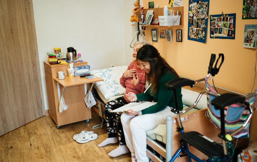 Pečovatelka sedí na posteli se seniorkou a společně se smějí