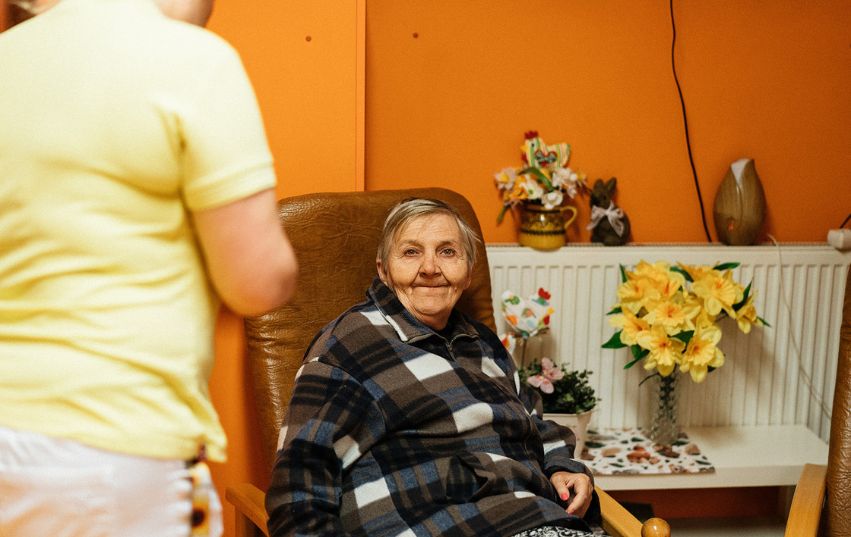 Pečovatelka stojí vedle seniorky sedící v křesle