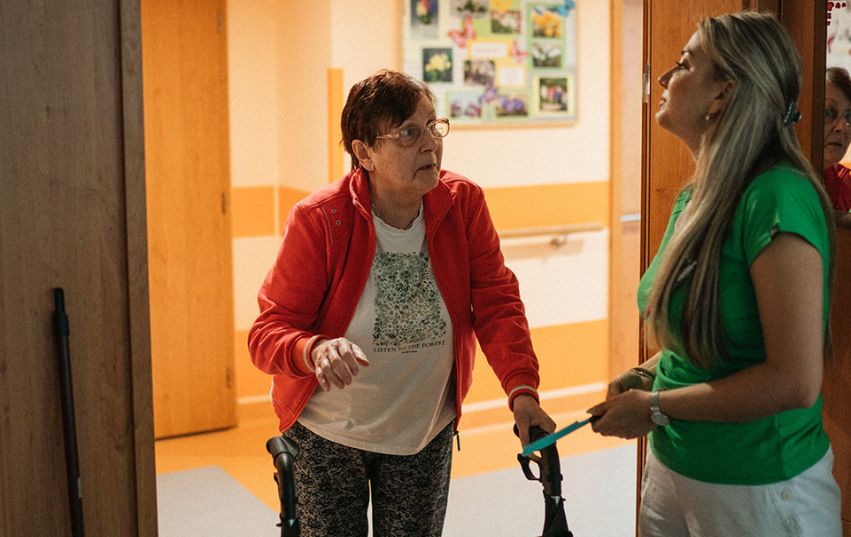 Seniorka s chod&iacute;tkem hovoř&iacute; s pečovatelkou na chodbě domova